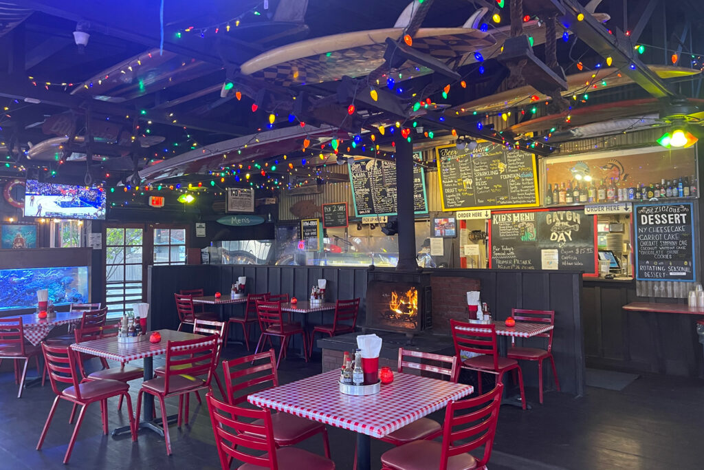 Interior photo of Reel Inn's indoor dining room with red gingham table clothes on the dining tables. Surfboards hang from the ceiling which has muticolored strands of Christmas lights hanging from the rafters.